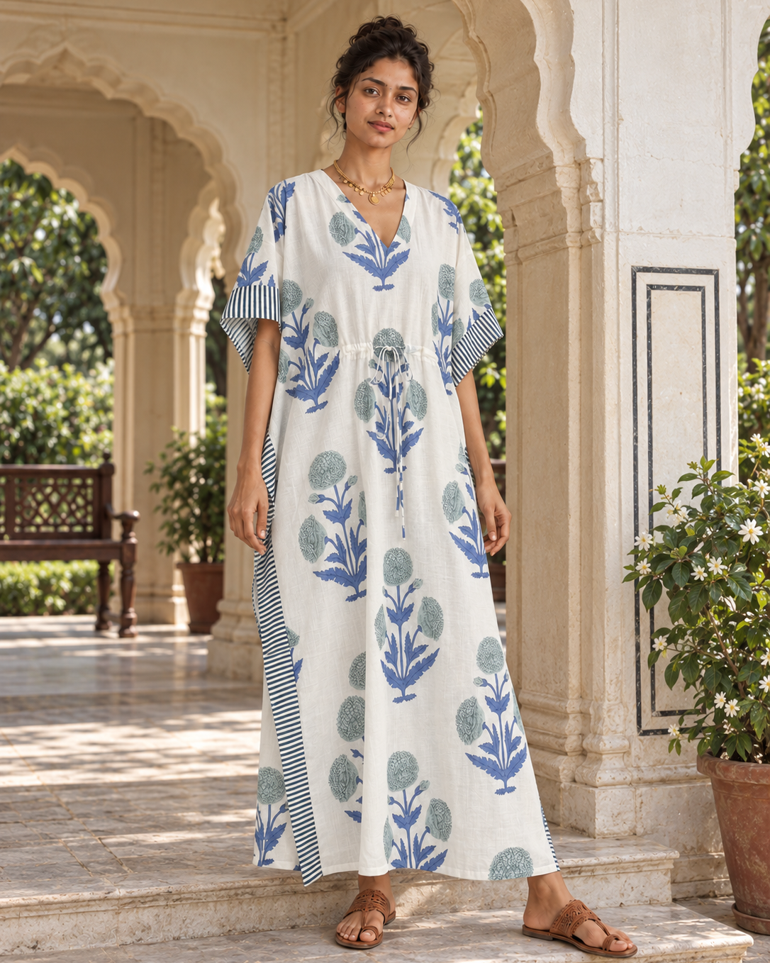 A woman stands on stone steps in an arched courtyard wearing a loose white kaftan dress with blue botanical prints and striped sleeve panels, paired with brown sandals.