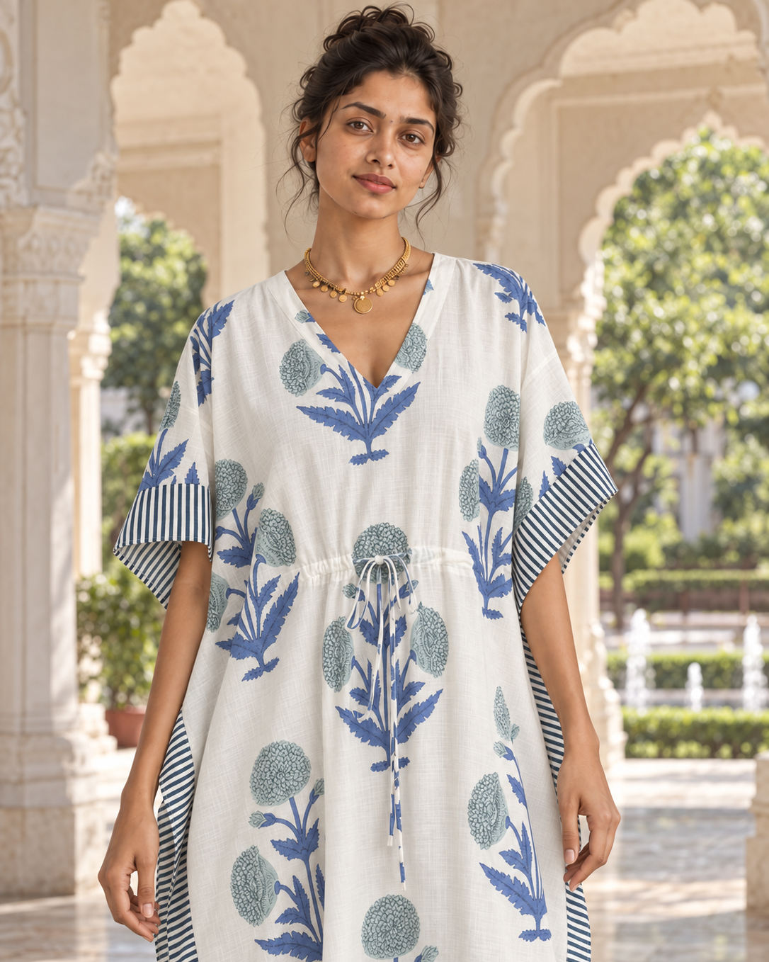 A woman wearing a white kaftan dress with blue floral block prints and striped sleeve/collar details, standing in an outdoor architectural setting with arches.