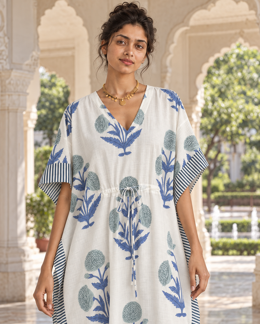 A woman wearing a white kaftan dress with blue floral block prints and striped sleeve/collar details, standing in an outdoor architectural setting with arches.