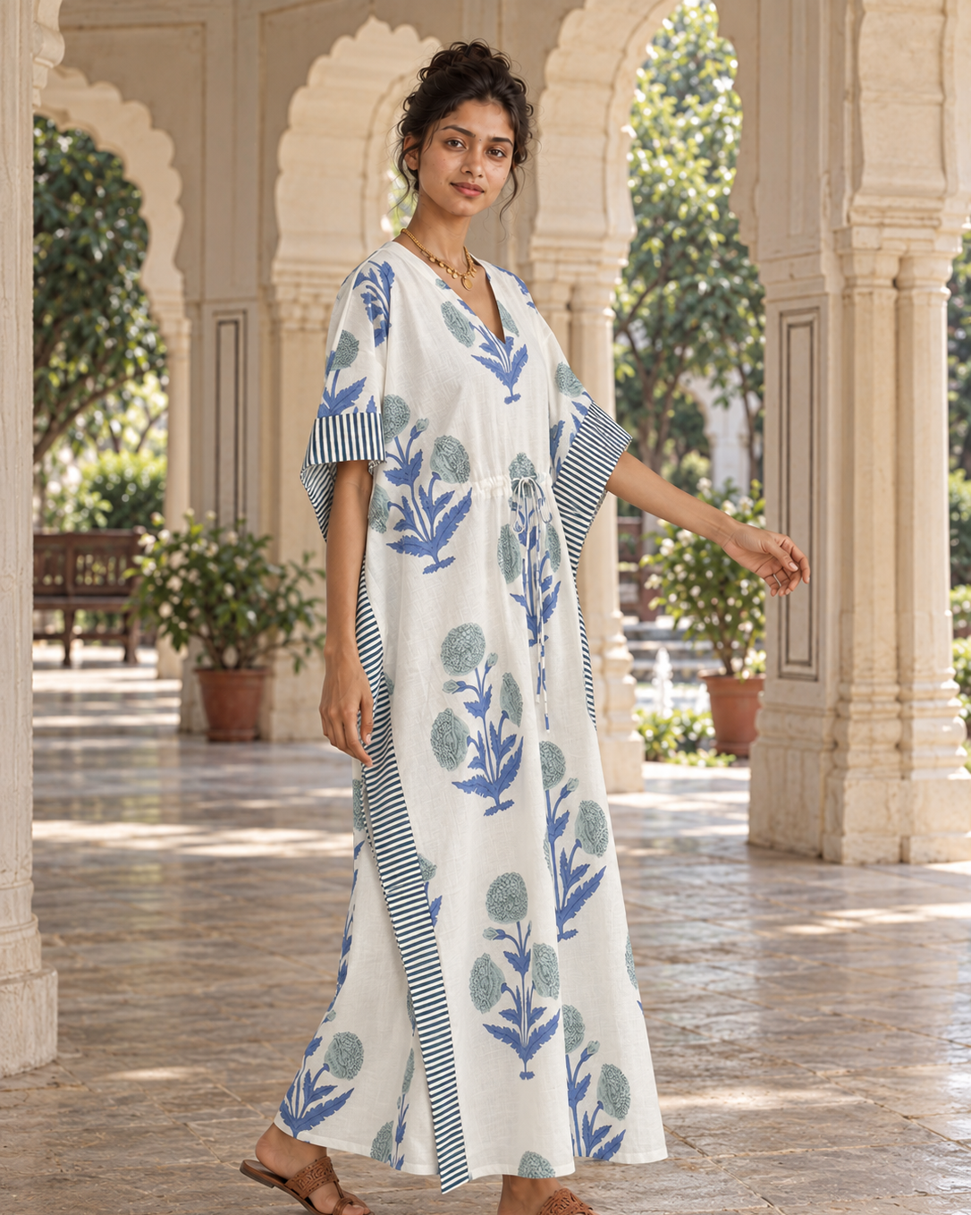 A woman models a white kaftan dress with blue floral and striped patterns in a traditional courtyard with arches.