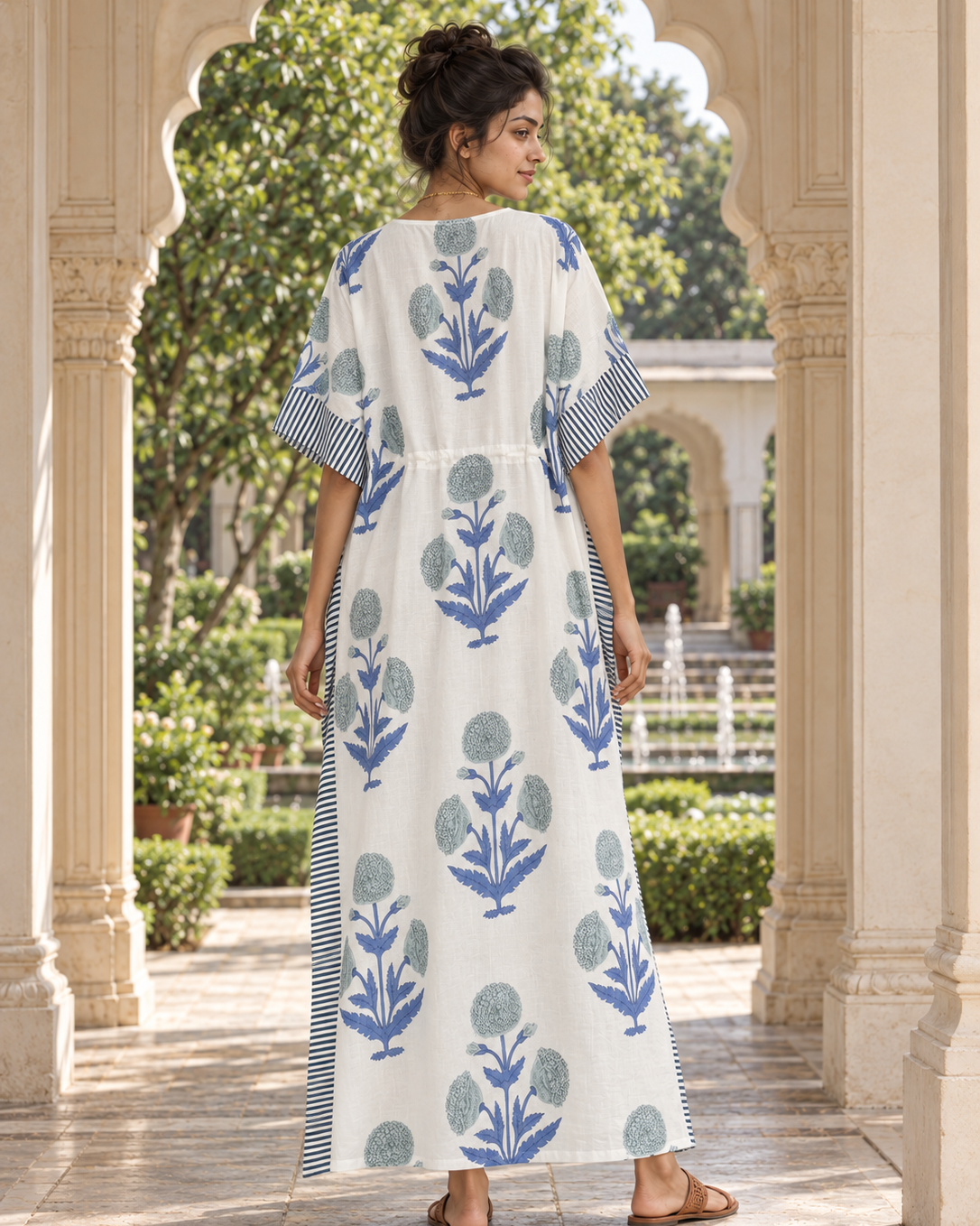 A woman stands in a courtyard under arched stone columns, wearing a long white kaftan dress with blue floral block prints and striped sleeve and side details.