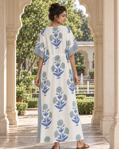A woman stands in a courtyard under arched stone columns, wearing a long white kaftan dress with blue floral block prints and striped sleeve and side details.